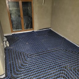 PanHydraulik - Niebieskie rury ogrzewania podłogowego ułożone na czarnej macie w surowym pomieszczeniu, widoczne przewody elektryczne i okno z folią.