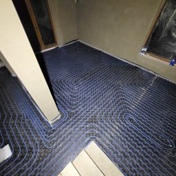 PanHydraulik - Niebieskie rurki ogrzewania podłogowego ułożone na czarnej macie w surowym wnętrzu. Widoczne okna zabezpieczone folią i elementy konstrukcyjne pomieszczenia.