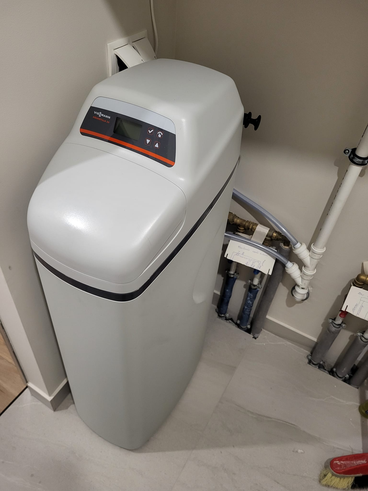 Zamontowany zmiękczacz wody Viessmann Aquastilla 25 z podłączonymi rurami w narożniku pomieszczenia z szarą podłogą i beżowymi ścianami. Widoczne schematy instalacji.