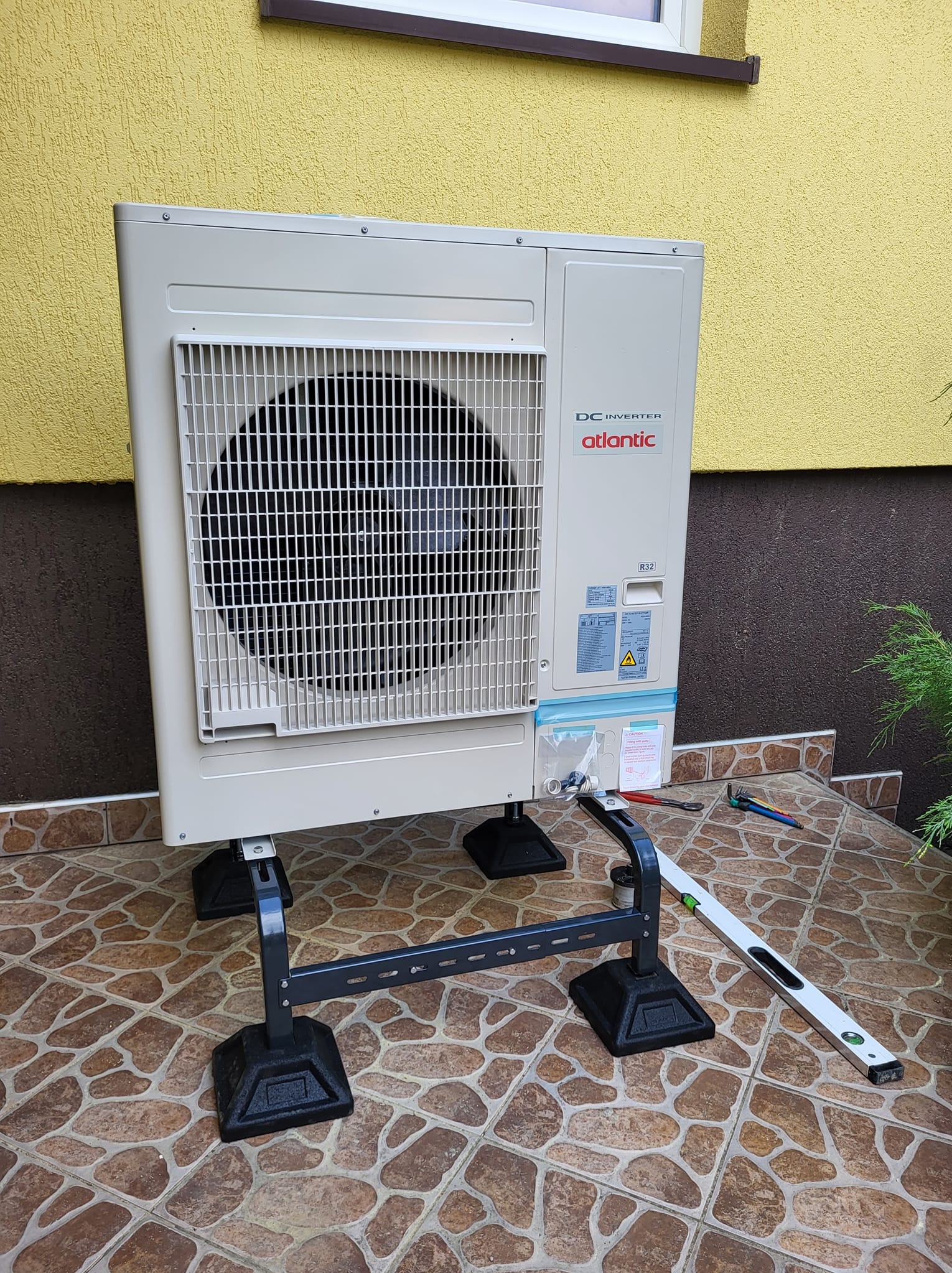 Biała jednostka zewnętrzna klimatyzacji Atlantic DC Inverter, zamontowana na czarnej konstrukcji wsporczej, poziomica obok, tło: żółta ściana i brązowa podłoga.