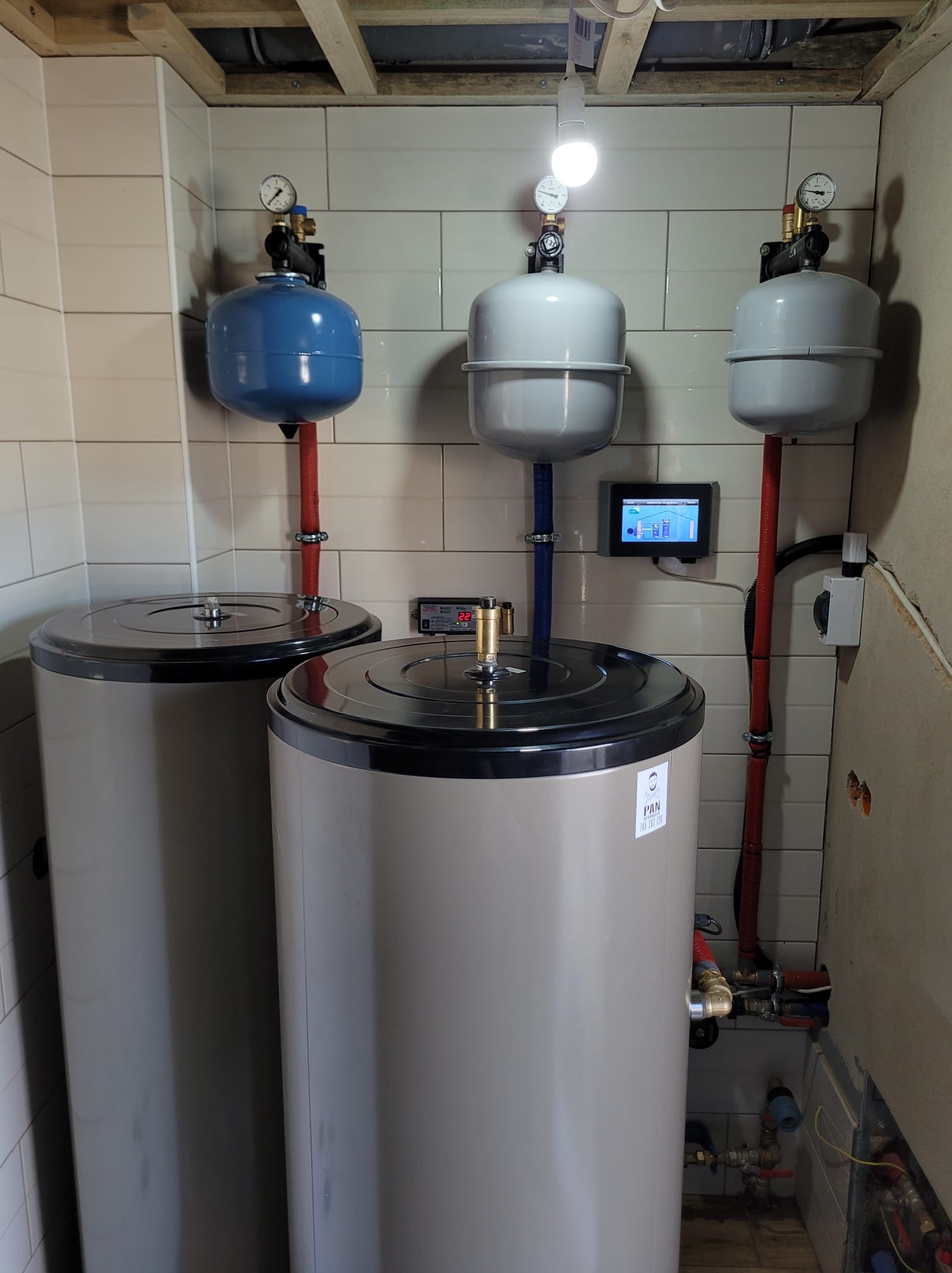 Trzy zbiorniki buforowe z osprzętem w pomieszczeniu z wykończonymi płytkami i widoczną instalacją elektryczną. Widoczny panel sterowania i manometry.