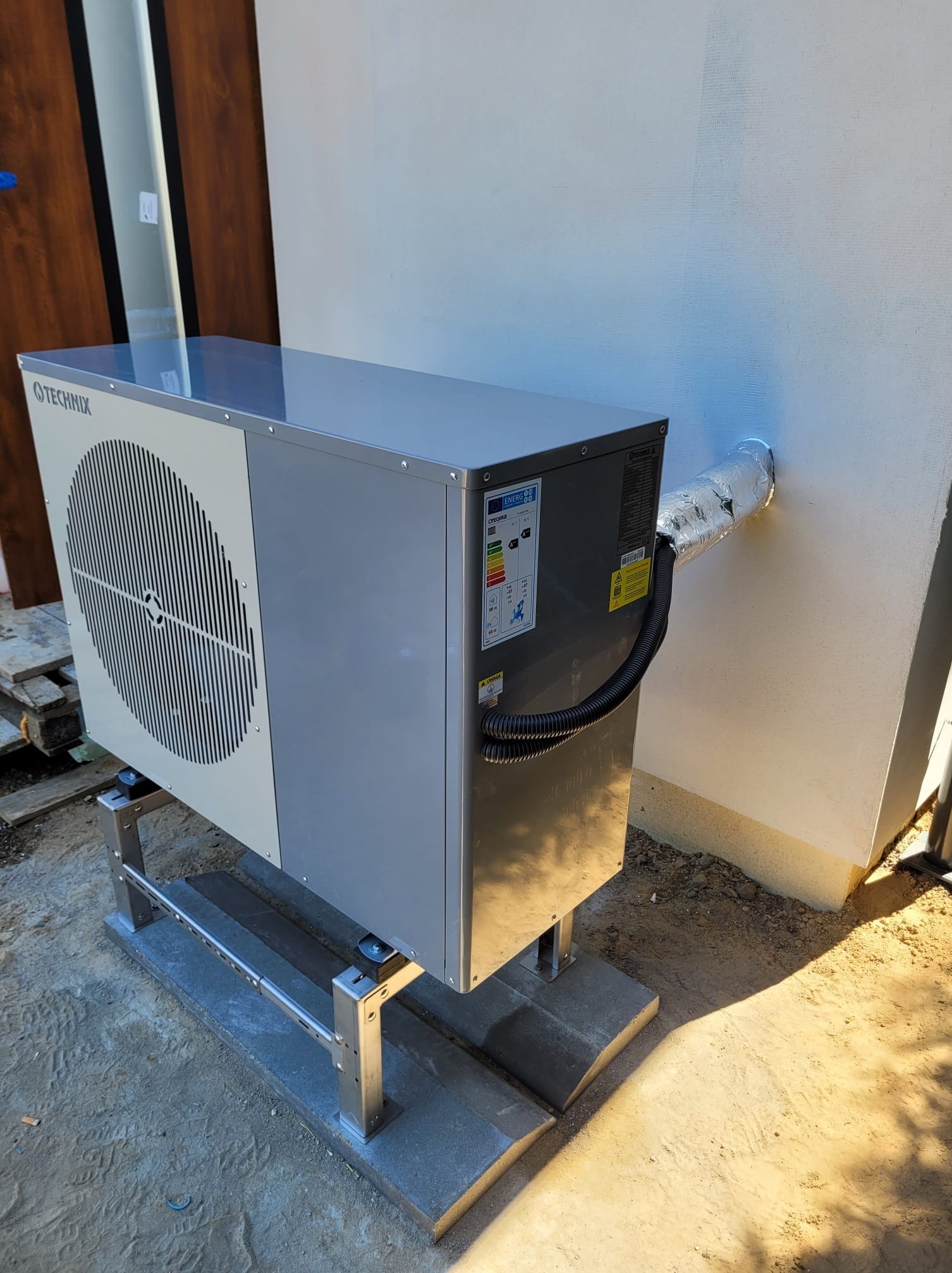 Szara pompa ciepła Technix zamontowana na metalowej konstrukcji, z podłączeniem do ściany budynku. Widoczna etykieta energetyczna.