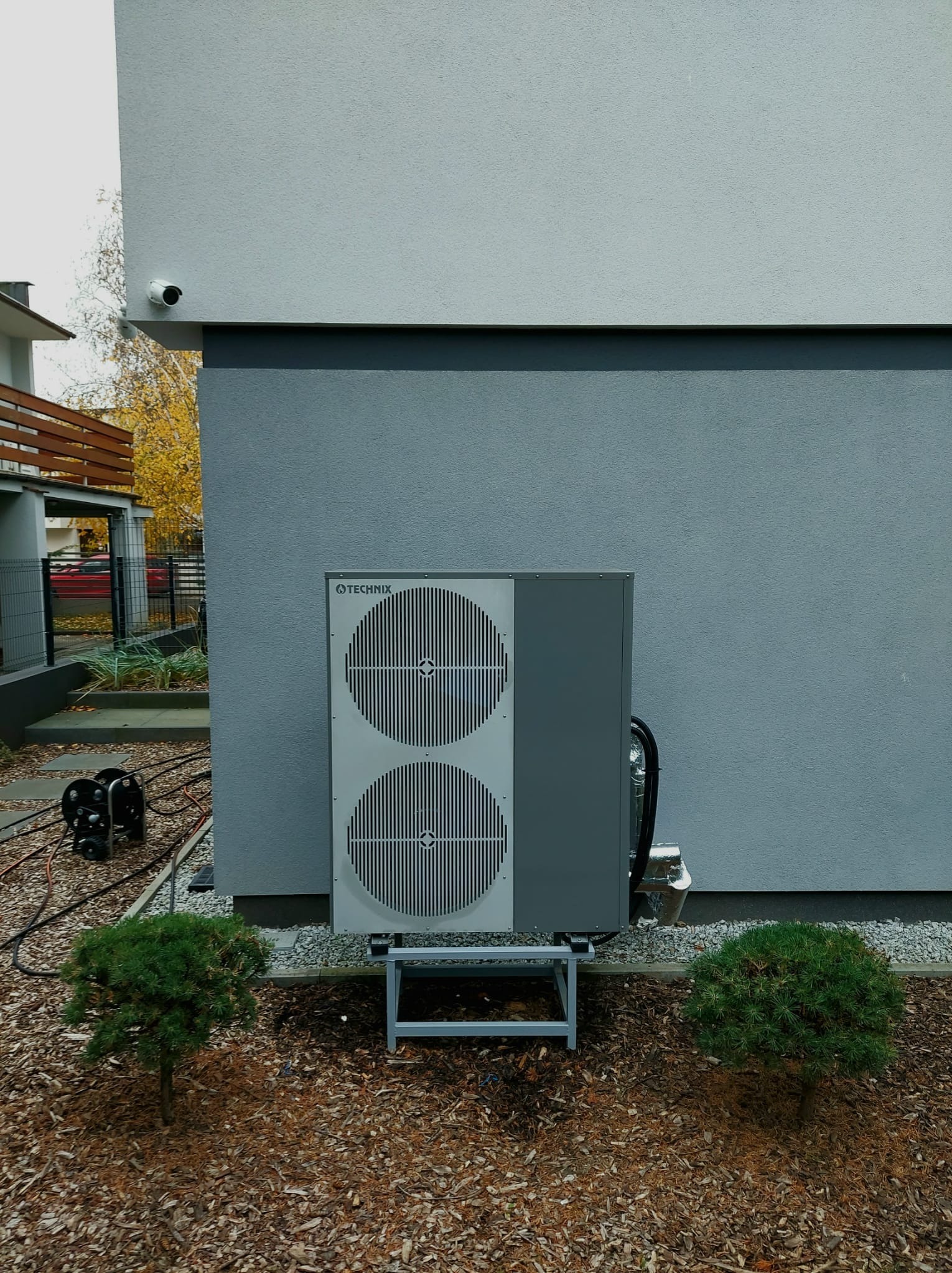 Szara pompa ciepła Technix zamontowana na metalowym stelażu, przy ścianie budynku z kamerą monitoringu. Obok małe drzewka iglaste.