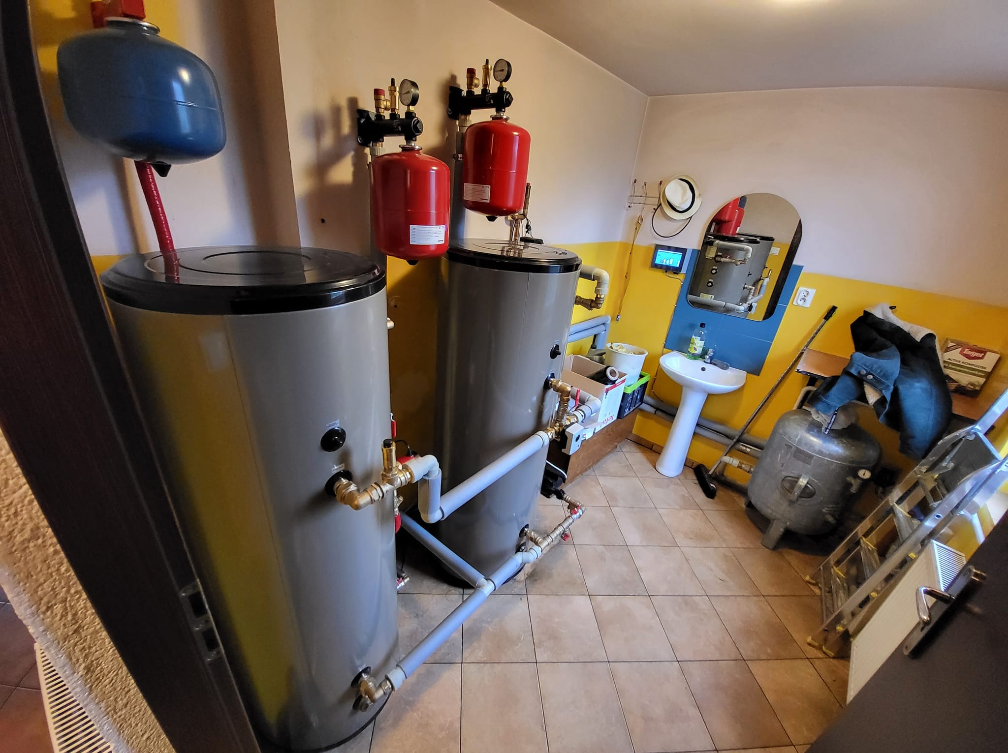 Dwa szare bojlery z czerwonymi zbiornikami wyrównawczymi połączone rurami w kotłowni; widoczna umywalka i drabina. Nietypowe żółte i białe ściany.