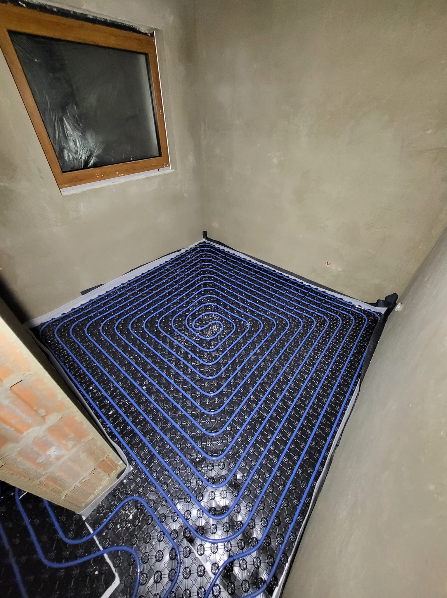 Niebieskie rury ogrzewania podłogowego ułożone spiralnie na czarnej macie w surowym pomieszczeniu z oknem i fragmentem cegły. Instalacja przed wylaniem posadzki.