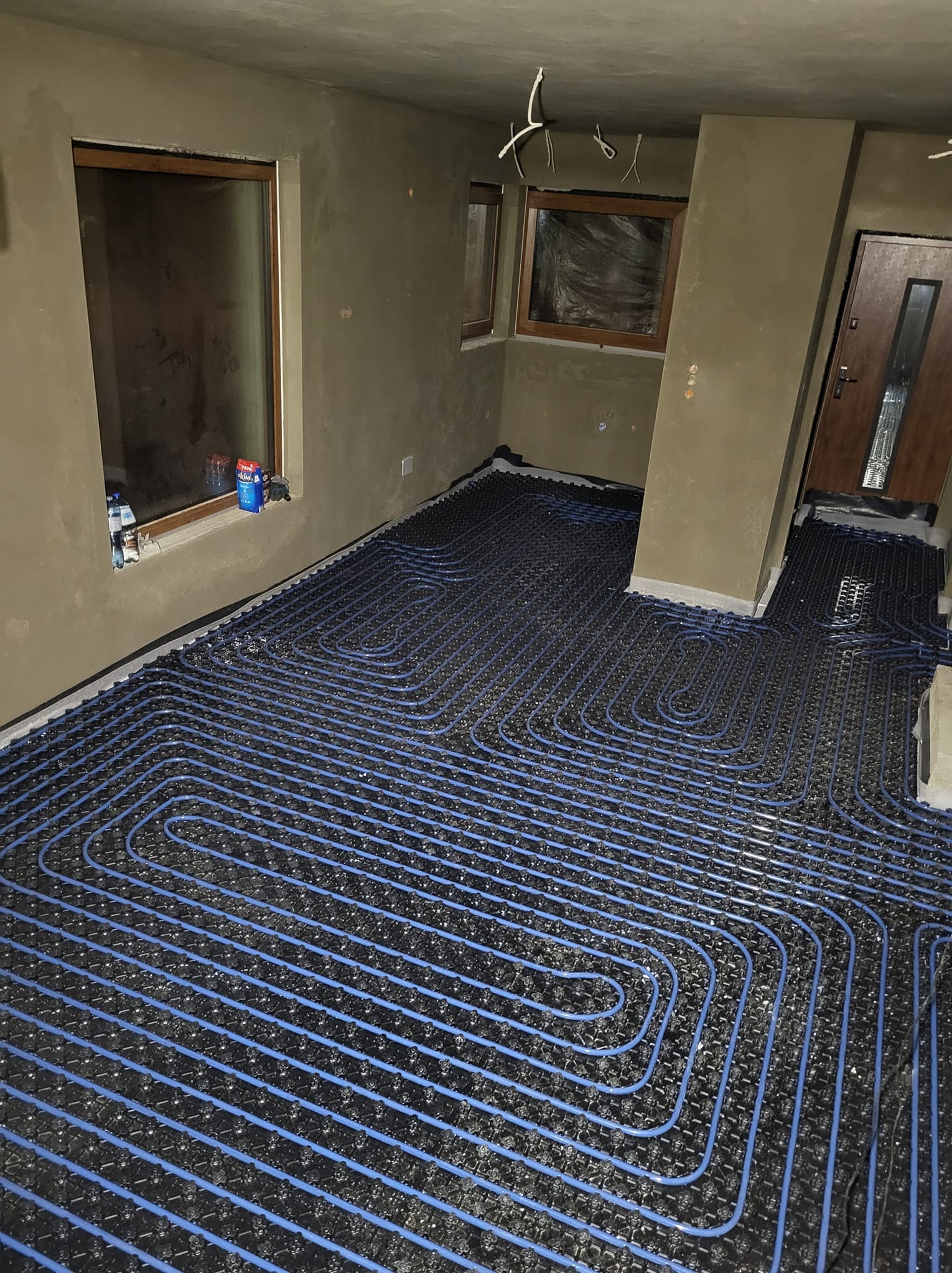 Niebieskie rury ogrzewania podłogowego ułożone spiralnie na czarnej macie w surowym wnętrzu z oknami i drzwiami, gotowe do wylania posadzki.