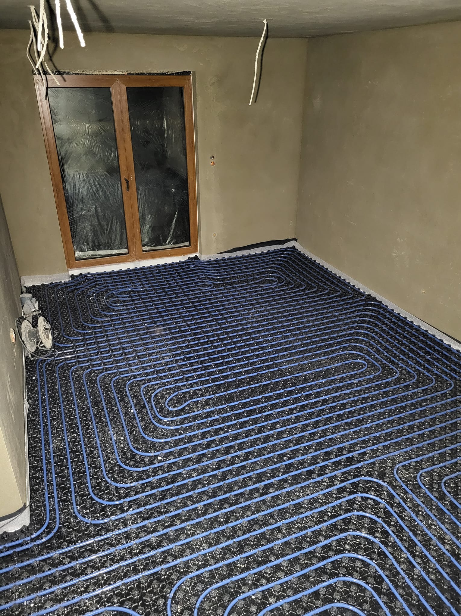 Niebieskie rury ogrzewania podłogowego ułożone na czarnej macie w surowym pomieszczeniu, widoczne przewody elektryczne i okno z folią.