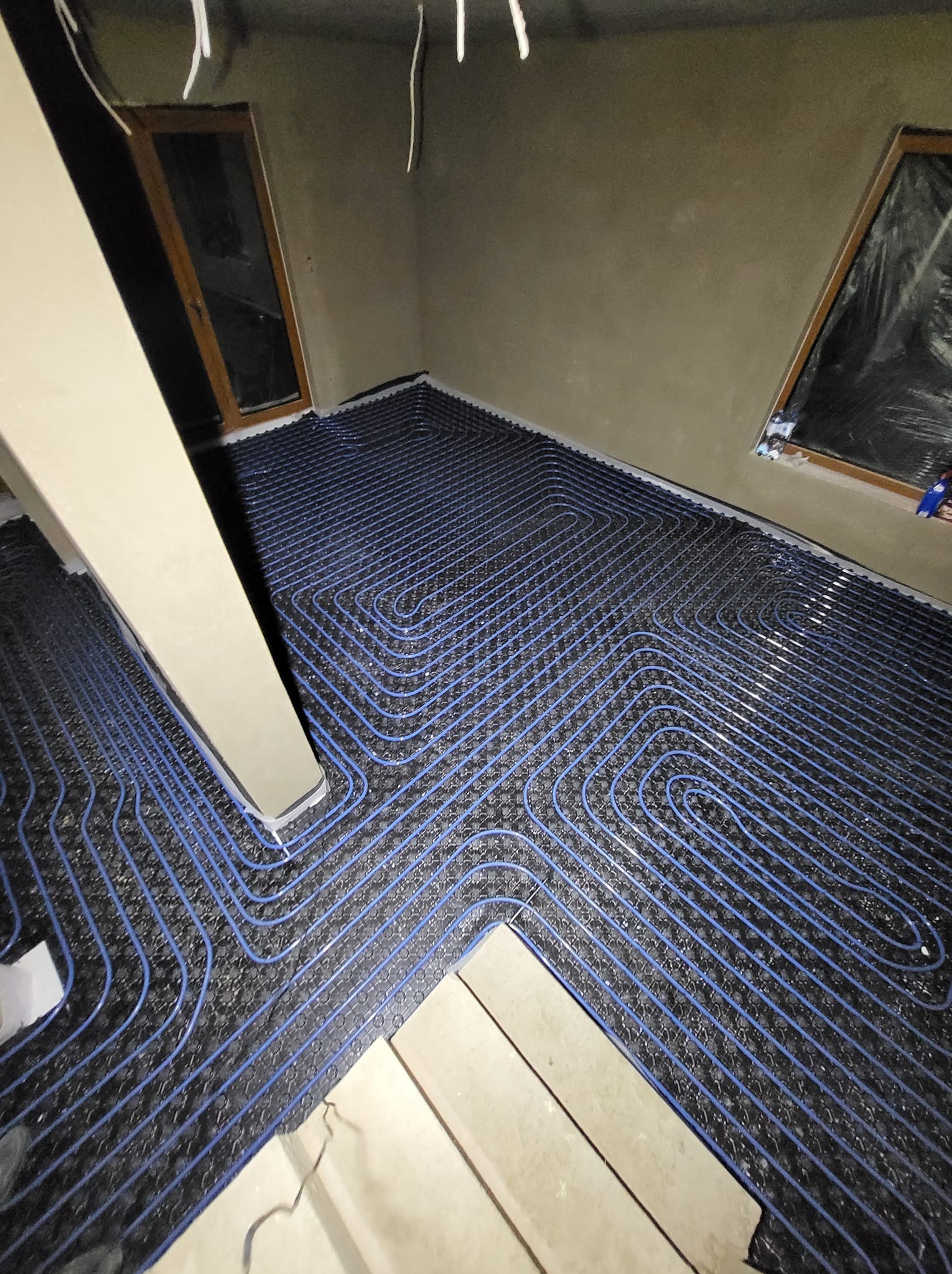 Niebieskie rurki ogrzewania podłogowego ułożone na czarnej macie w surowym wnętrzu. Widoczne okna zabezpieczone folią i elementy konstrukcyjne pomieszczenia.