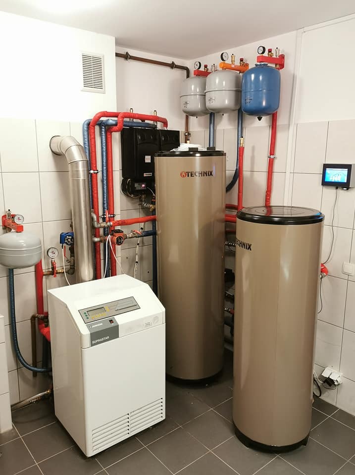 Kompletna kotłownia: dwa bojlery Technix, biały piec Supastar, czarny moduł sterujący, naczynia przeponowe i rury w pomieszczeniu wyłożonym płytkami.