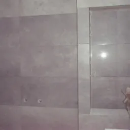 Ściana wyłożona szarymi płytkami w łazience w Jeleniej Górze, z dwoma wystającymi elementami instalacji i wnęką, w której stoi kartonowe pudełko.