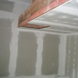 Montaż płyty karton gips na suficie podwieszanym z widoczną drewnianą konstrukcją nośną i niedokończonymi ścianami w surowym stanie.