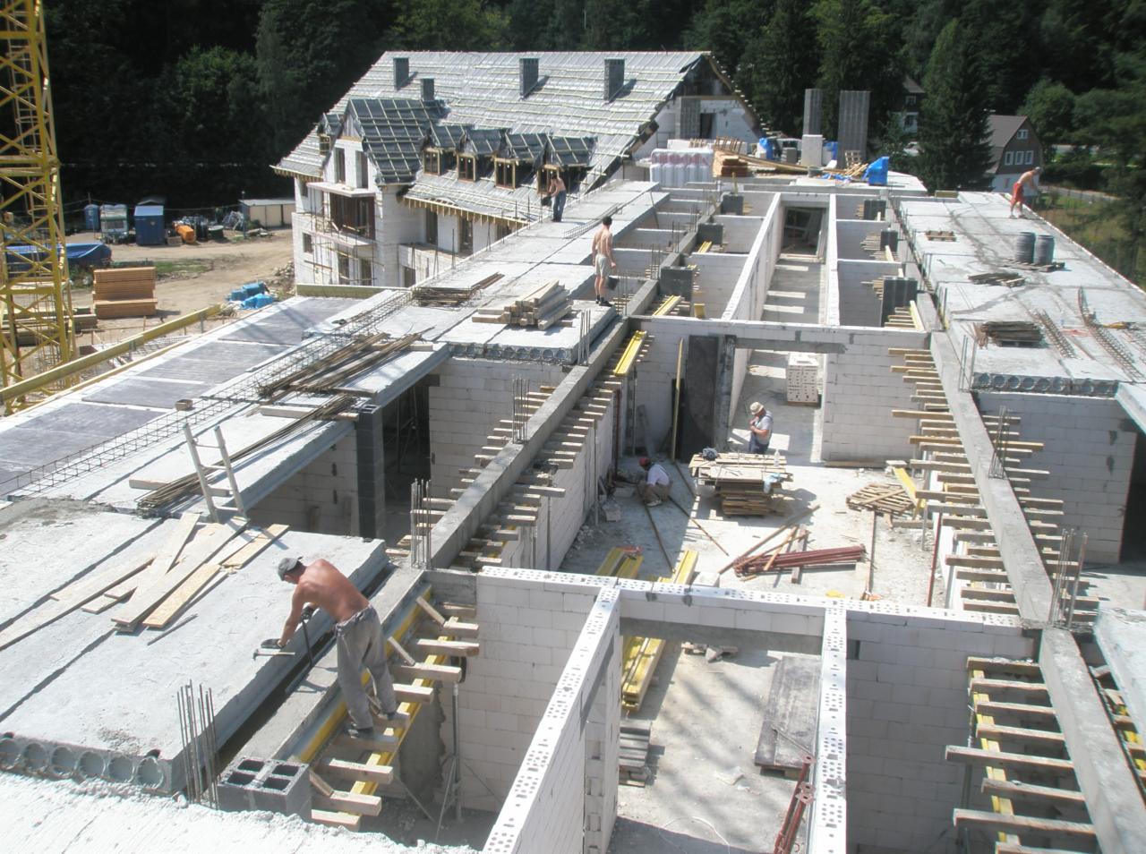 Wysoki kąt widzenia na budowę domu w stanie surowym otwartym, widoczne ściany z bloczków, betonowe stropy, zbrojenia, drewniane szalunki oraz pracownicy na budowie.