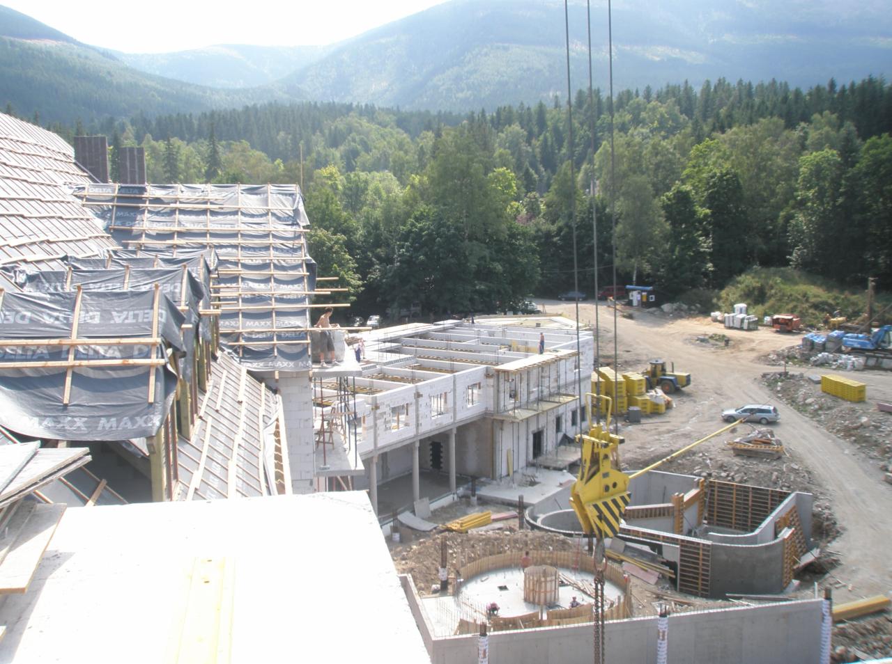 Budowa domu w stanie surowym otwartym z widocznymi elementami konstrukcyjnymi dachu i ścian, na tle zalesionych gór.