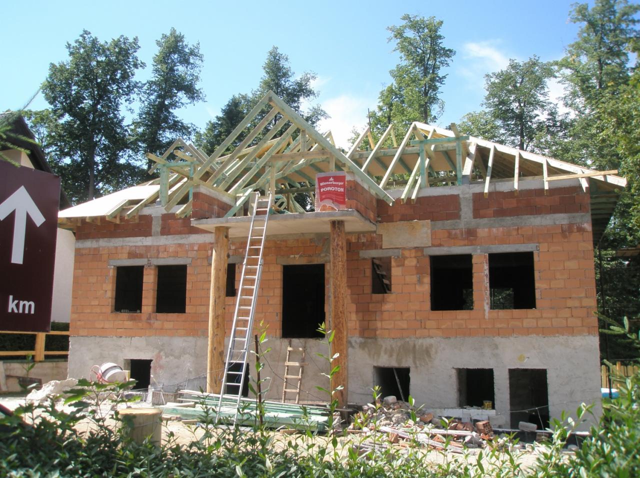 Budowa domu jednorodzinnego w stanie surowym otwartym z widoczną konstrukcją dachu, murem z czerwonej cegły Porotherm i drabina oparta o ścianę.