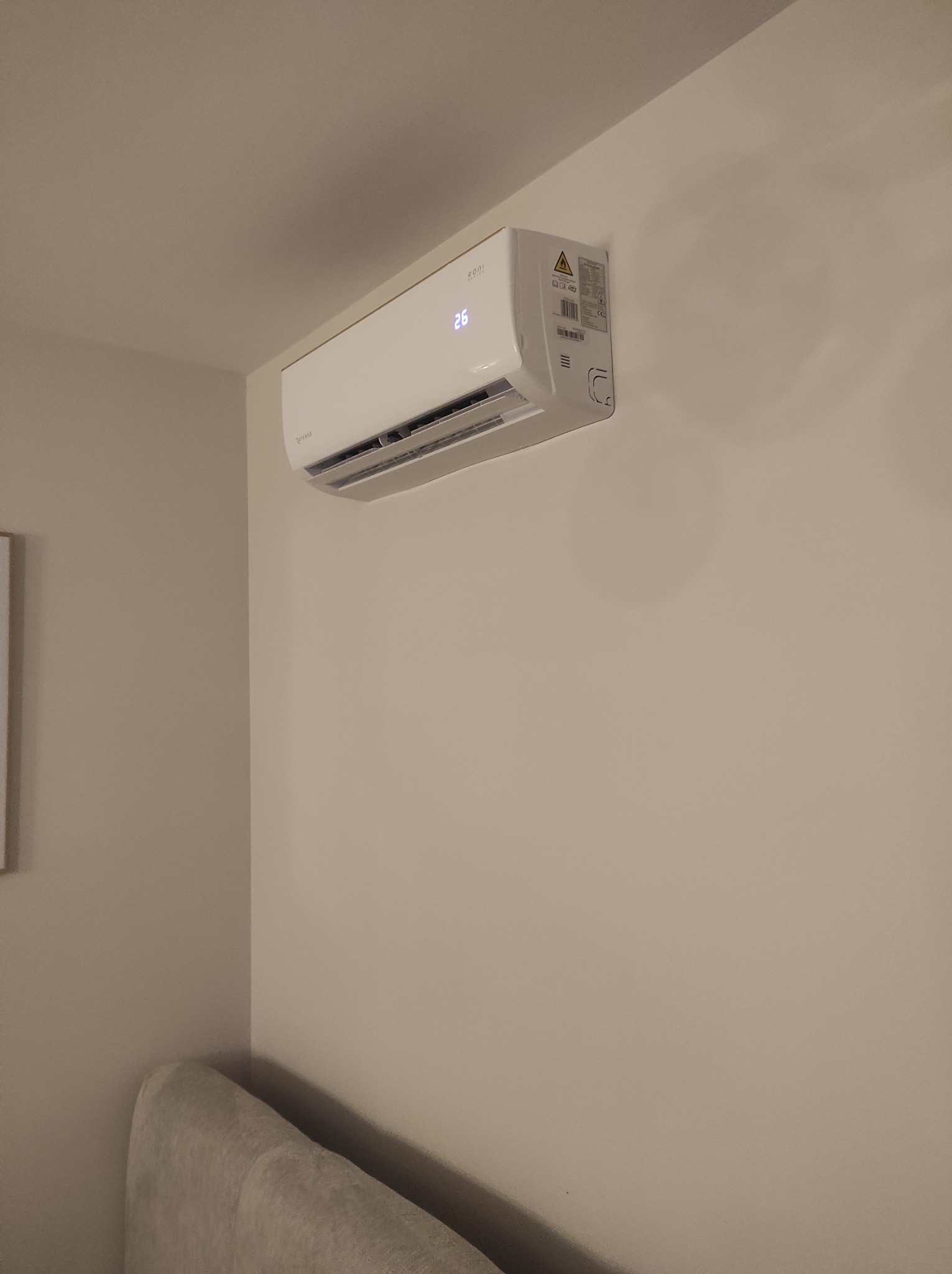 Biały klimatyzator ścienny z wyświetlaczem temperatury 26 stopni, zamontowany w jasnym pomieszczeniu. Widoczna część sofy.