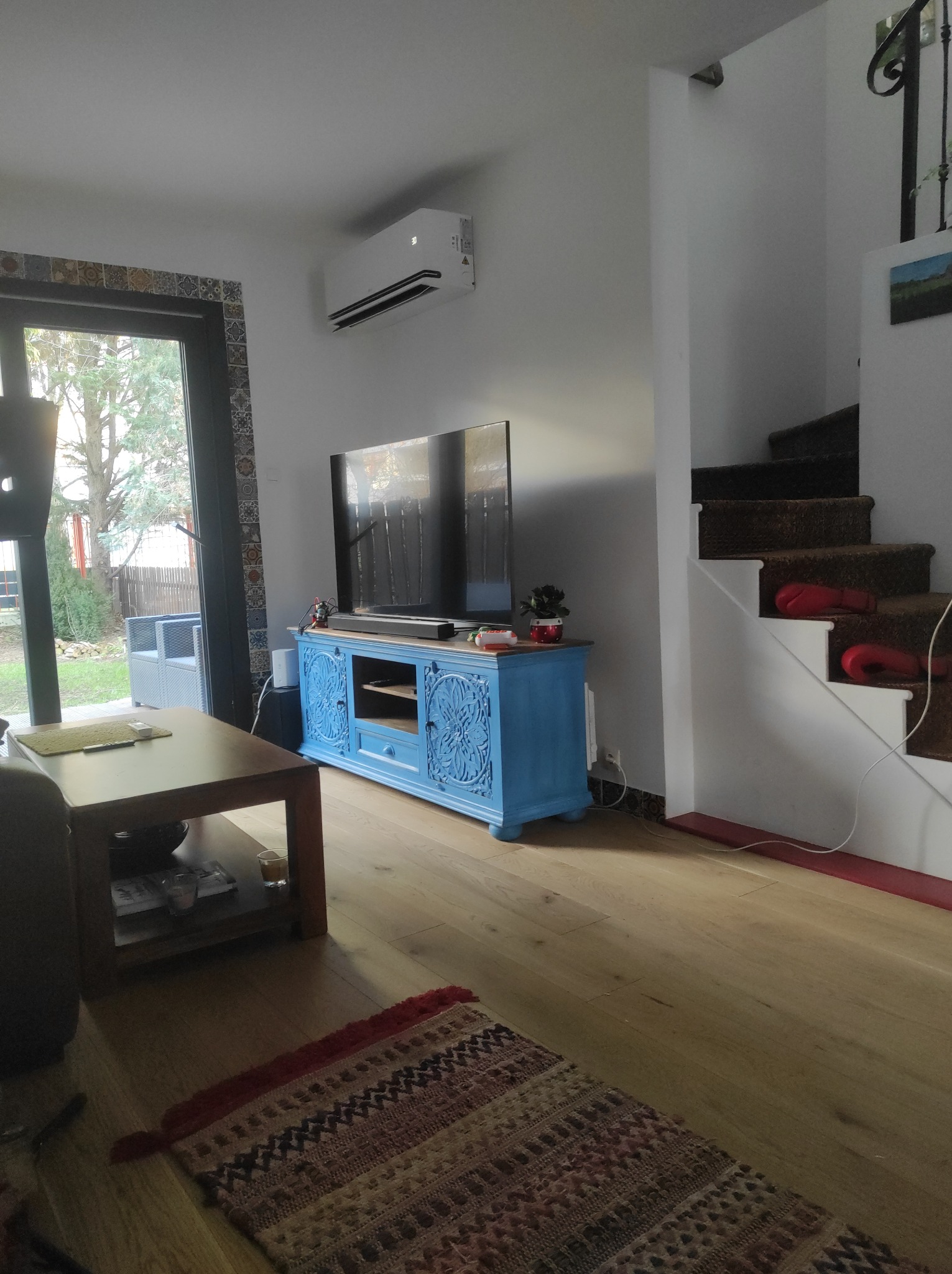 Biała klimatyzacja ścienna w salonie z niebieską komodą pod telewizorem, drewnianą podłogą i schodami. Widok na ogród przez przeszklone drzwi.