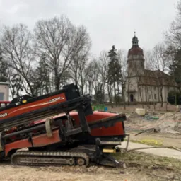 Ditch Witch JT3020 w trakcie przewiertu sterowanego, na tle kościoła i drzew, w pochmurny dzień. Widoczny sprzęt do wiercenia horyzontalnego.