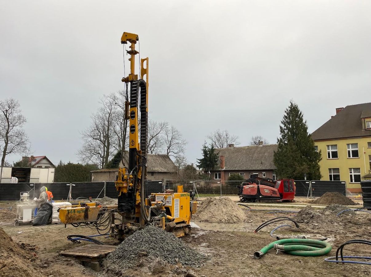 Żółta wiertnica Beretta T44 na placu budowy, z pracownikiem w kombinezonie i czerwoną maszyną w tle, pod pochmurnym niebem. Widoczny gruz i elementy instalacji.