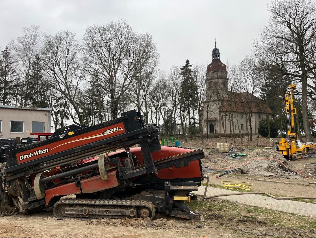 Ditch Witch JT3020 w trakcie przewiertu sterowanego, na tle kościoła i drzew, w pochmurny dzień. Widoczny sprzęt do wiercenia horyzontalnego.
