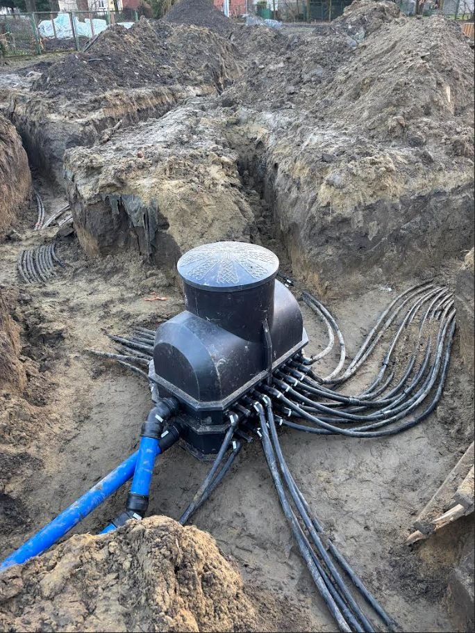 Rozdzielnica w wykopie z licznymi kablami i rurami, teren budowy z ziemią w tle. Instalacja podziemna z widocznymi elementami przyłączeniowymi.