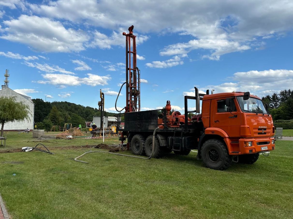 Pomarańczowa wiertnica KAMAZ na trawniku, w tle budynek i drzewa. Widoczne narzędzia i etap pracy. Błękitne niebo z obłokami.