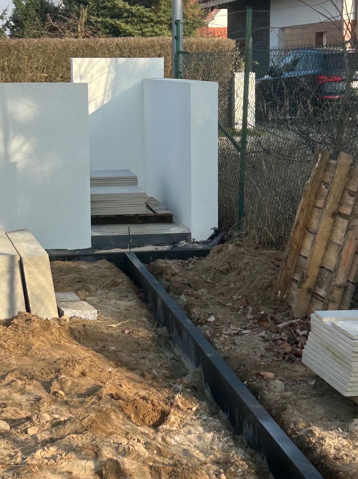 Budowa schodów zewnętrznych z jasnych płyt, widoczne elementy konstrukcyjne i przygotowany teren. Czarna listwa krawężnikowa oddziela teren.