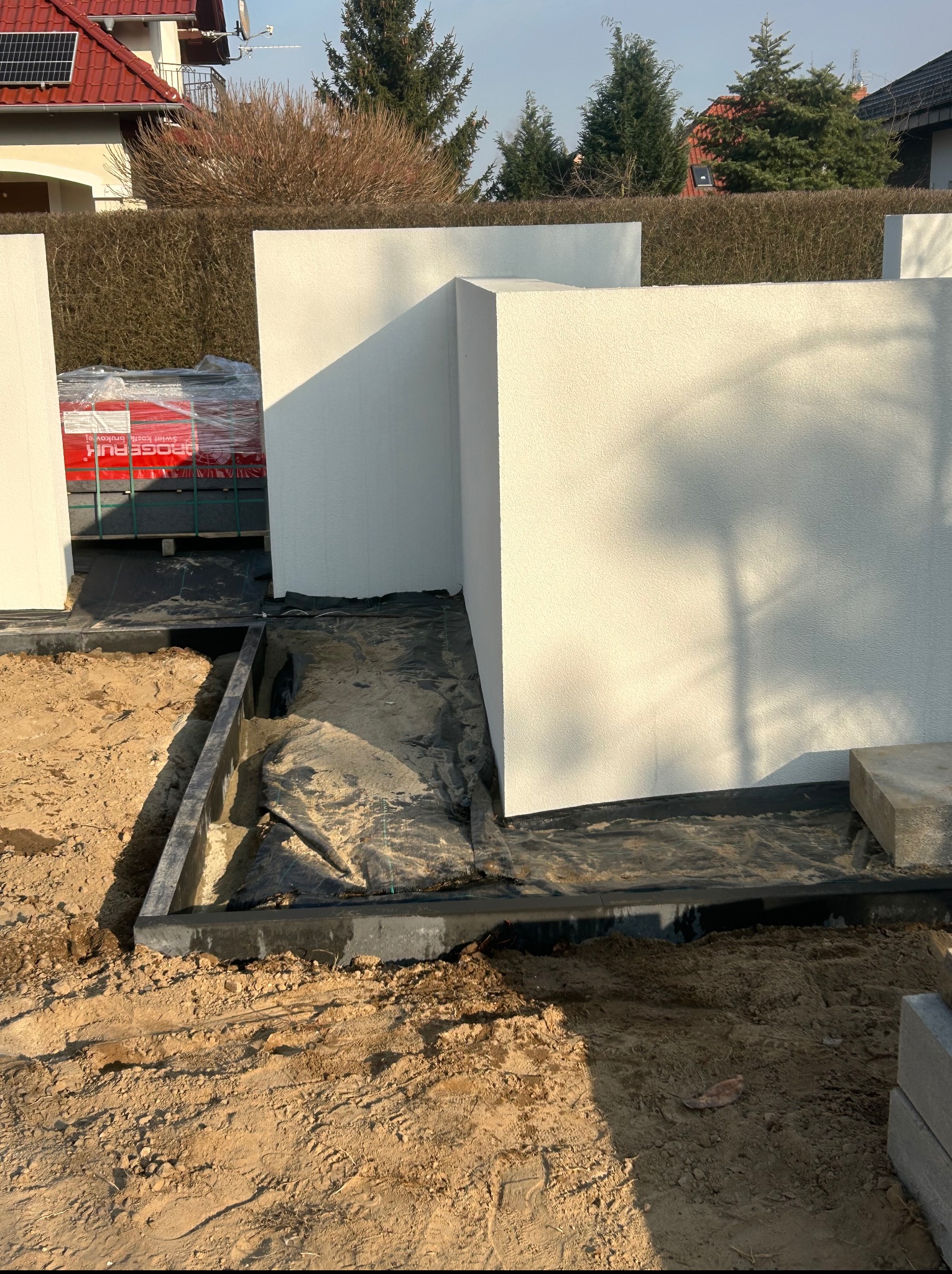 Budowa ogrodzenia z białych bloczków, widoczne fundamenty i materiały budowlane. W tle żywopłot i dom z czerwoną dachówką.