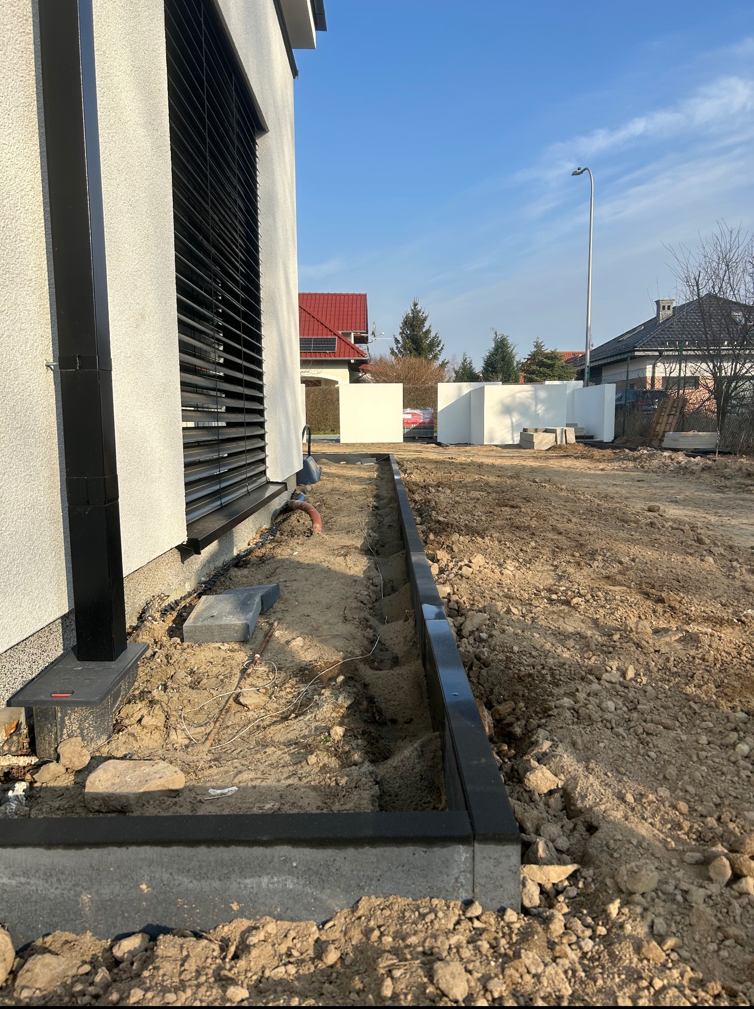 Przygotowanie terenu pod obrzeża rabatki przy nowoczesnym domu z czarnymi elementami wykończenia. Widoczne wykopy i materiały budowlane.