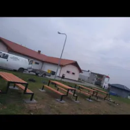 Rząd metalowo-drewnianych ławek na trawiastym terenie, w tle budynki i zaparkowany biały van. Nietypowy kształt nóg ławek, pochmurne niebo.