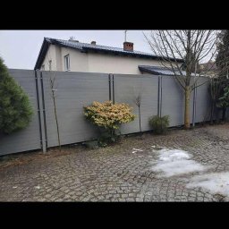 Kamil Aszyk K.G.J.W - Nowoczesne ogrodzenie z szarych paneli kompozytowych, oddzielające posesję od ulicy. Widoczne krzewy i drzewa wzdłuż ogrodzenia, kostka brukowa na podjeździe.