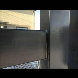 Kamil Aszyk K.G.J.W - Detal metalowej konstrukcji ogrodzenia, spawane połączenie profili. Widoczne ślady po spawaniu, szlifowaniu i zabezpieczeniu antykorozyjnym. Tło rozmyte, budynek.