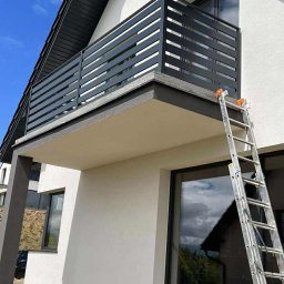 Kamil Aszyk K.G.J.W - Nowoczesna, pozioma balustrada balkonowa w kolorze antracytowym, z niskim murem, na tle białej elewacji domu i fragmentu drabiny oparta o ścianę budynku.