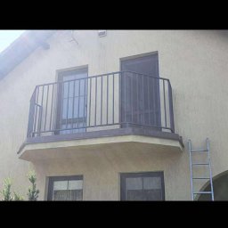 Kamil Aszyk K.G.J.W - Balkon z metalową, pionową balustradą w kolorze grafitowym. Widoczne drzwi balkonowe i okna. Z prawej strony drabina oparta o ścianę budynku.