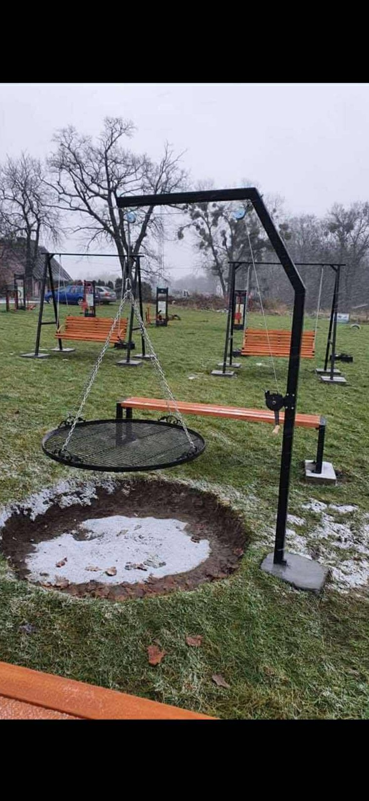 Metalowa konstrukcja z rusztem nad wykopanym dołem na ognisko, w tle huśtawki z drewnianymi siedziskami na trawiastym terenie. Widok w pochmurny dzień.