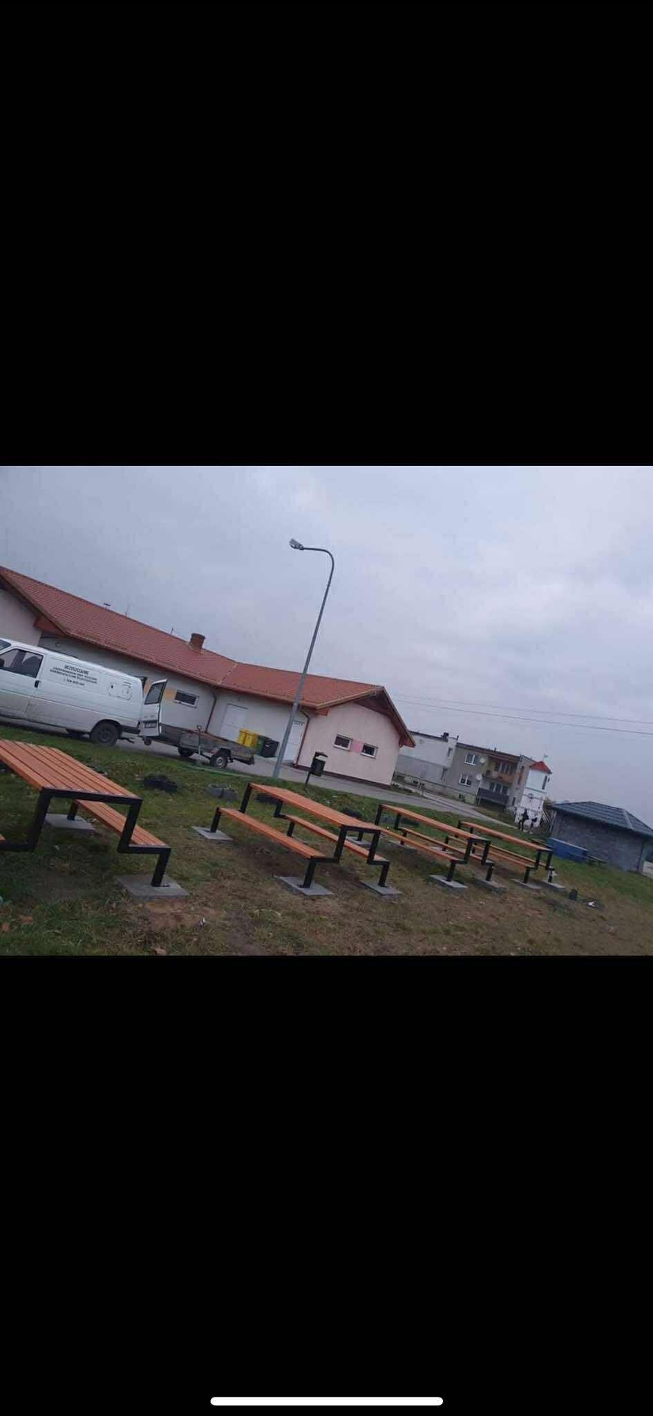 Rząd metalowo-drewnianych ławek na trawiastym terenie, w tle budynki i zaparkowany biały van. Nietypowy kształt nóg ławek, pochmurne niebo.