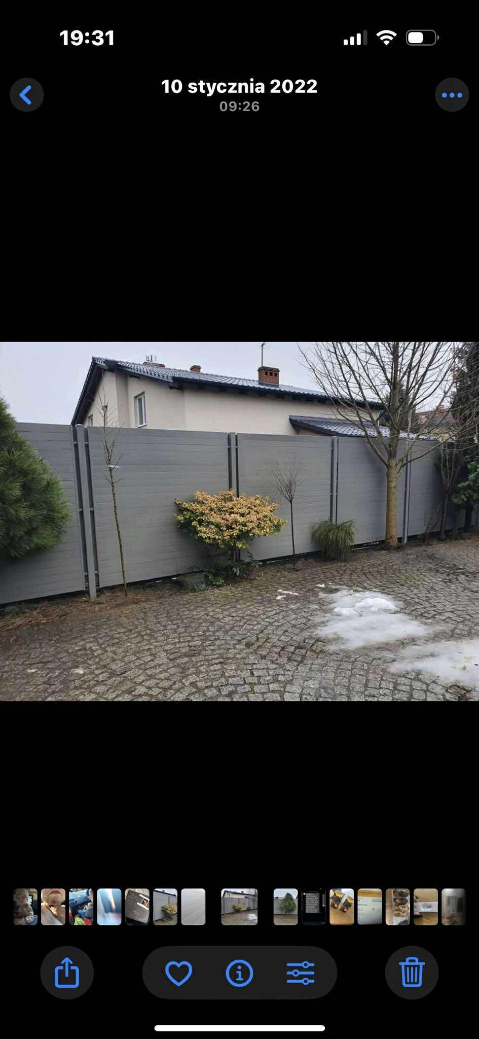 Nowoczesne ogrodzenie z szarych paneli kompozytowych, oddzielające posesję od ulicy. Widoczne krzewy i drzewa wzdłuż ogrodzenia, kostka brukowa na podjeździe.