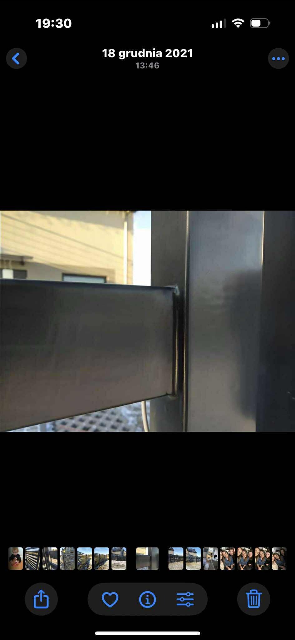 Detal metalowej konstrukcji ogrodzenia, spawane połączenie profili. Widoczne ślady po spawaniu, szlifowaniu i zabezpieczeniu antykorozyjnym. Tło rozmyte, budynek.