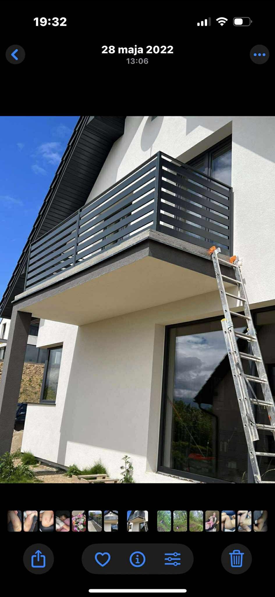 Nowoczesna, pozioma balustrada balkonowa w kolorze antracytowym, z niskim murem, na tle białej elewacji domu i fragmentu drabiny oparta o ścianę budynku.