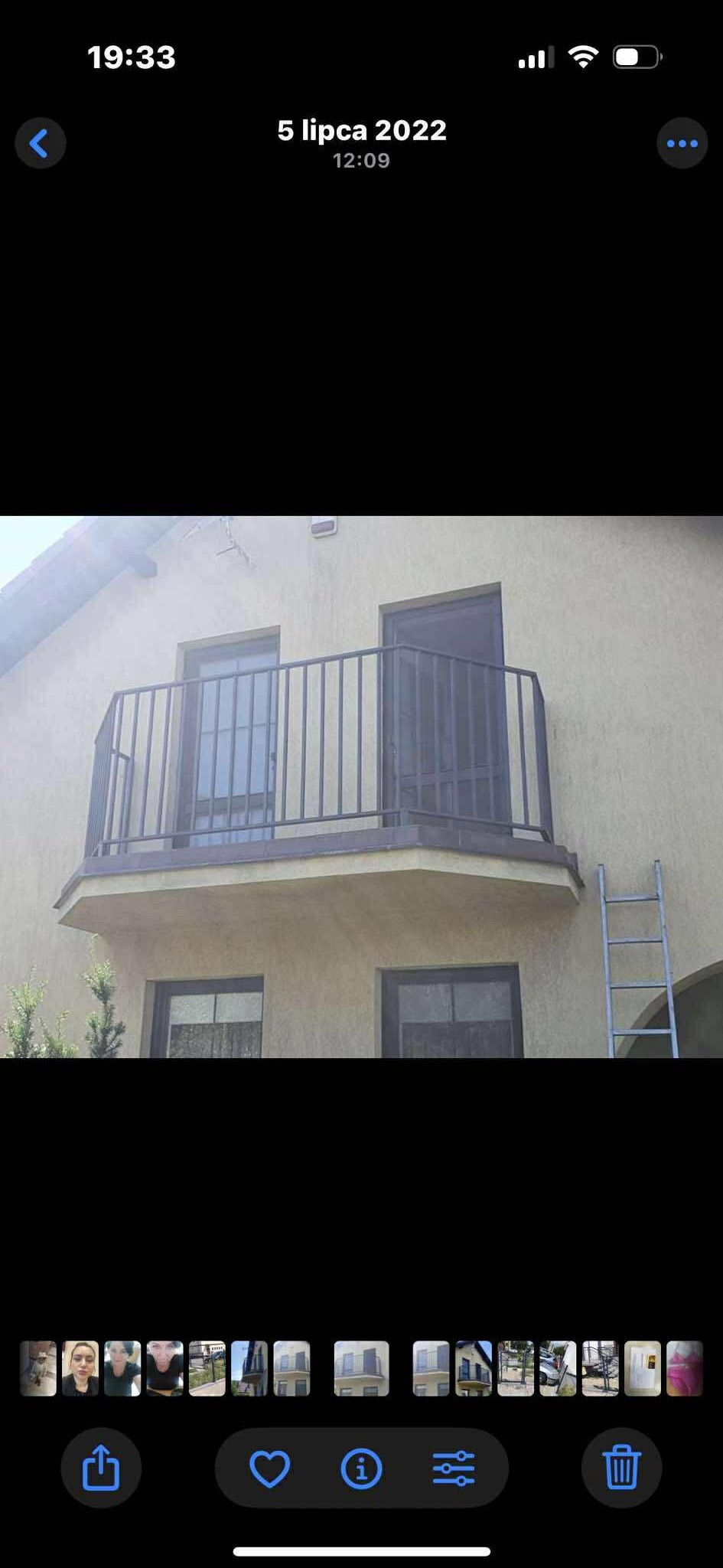 Balkon z metalową, pionową balustradą w kolorze grafitowym. Widoczne drzwi balkonowe i okna. Z prawej strony drabina oparta o ścianę budynku.