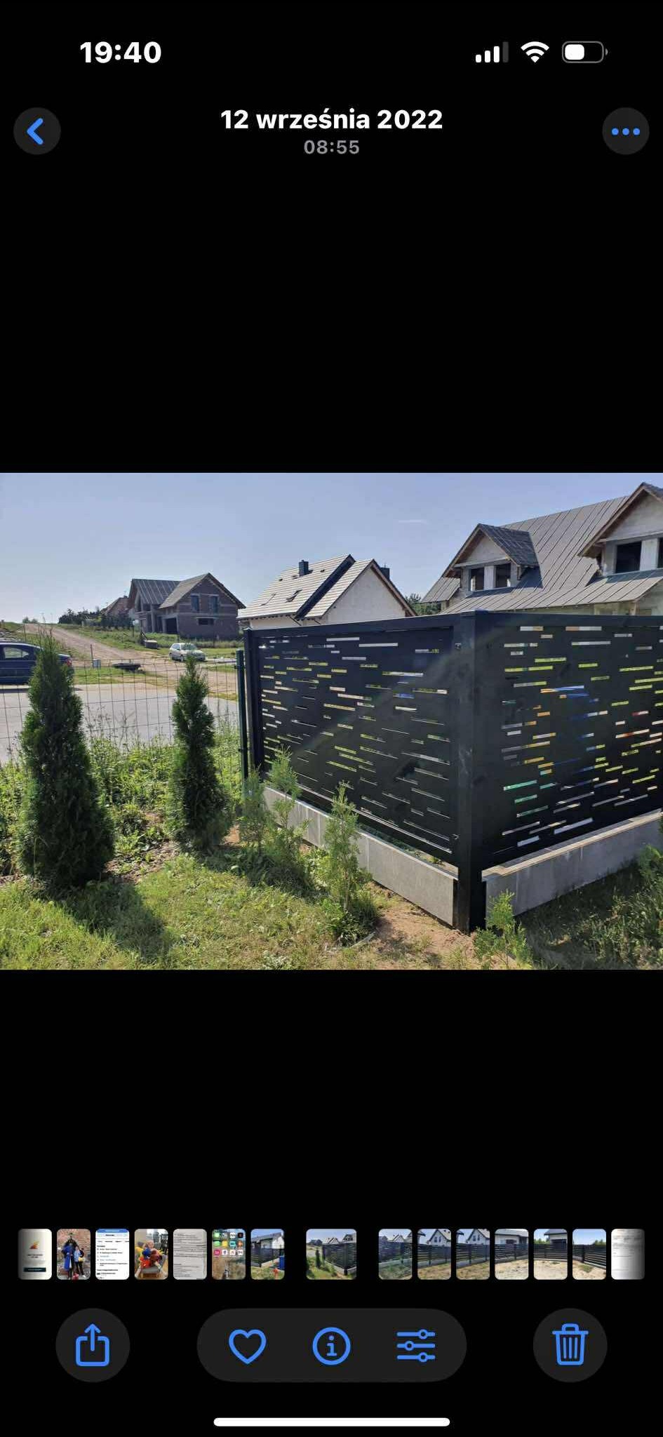 Nowoczesne, ażurowe ogrodzenie panelowe w kolorze grafitowym, z poziomymi szczelinami, na tle domów w budowie i zieleni. Widoczne tuje przy ogrodzeniu.