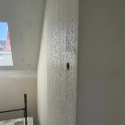 Ściana po nałożeniu dekoracyjnej struktury, widoczny fragment rusztowania i okno dachowe w skosie. Teksturowana powierzchnia ściany w jasnym odcieniu beżu.