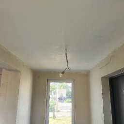 filar-BUD - Pokój po szpachlowaniu ścian i sufitu, przygotowany do dalszych prac wykończeniowych. Widoczne okno z widokiem na zieleń oraz wisząca żarówka.