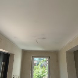 filar-BUD - Wnętrze pokoju po szpachlowaniu ścian i sufitu, widoczne wystające kable, okno z widokiem na zieleń. Przygotowanie do dalszych prac wykończeniowych.