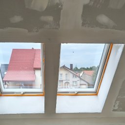 filar-BUD - Poddasze z dwoma oknami dachowymi po szpachlowaniu, widoczne ślady gładzi na ścianach i suficie. W oknach widok na domy z czerwonymi dachami.