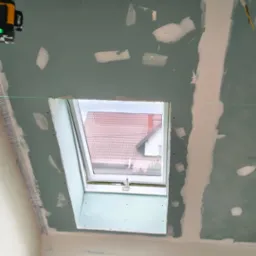 Montaż płyty KG na poddaszu z oknem dachowym. Widoczne szpachlowane łączenia, laser krzyżowy i fragment siatki na suficie. Surowy stan wykończenia wnętrza.