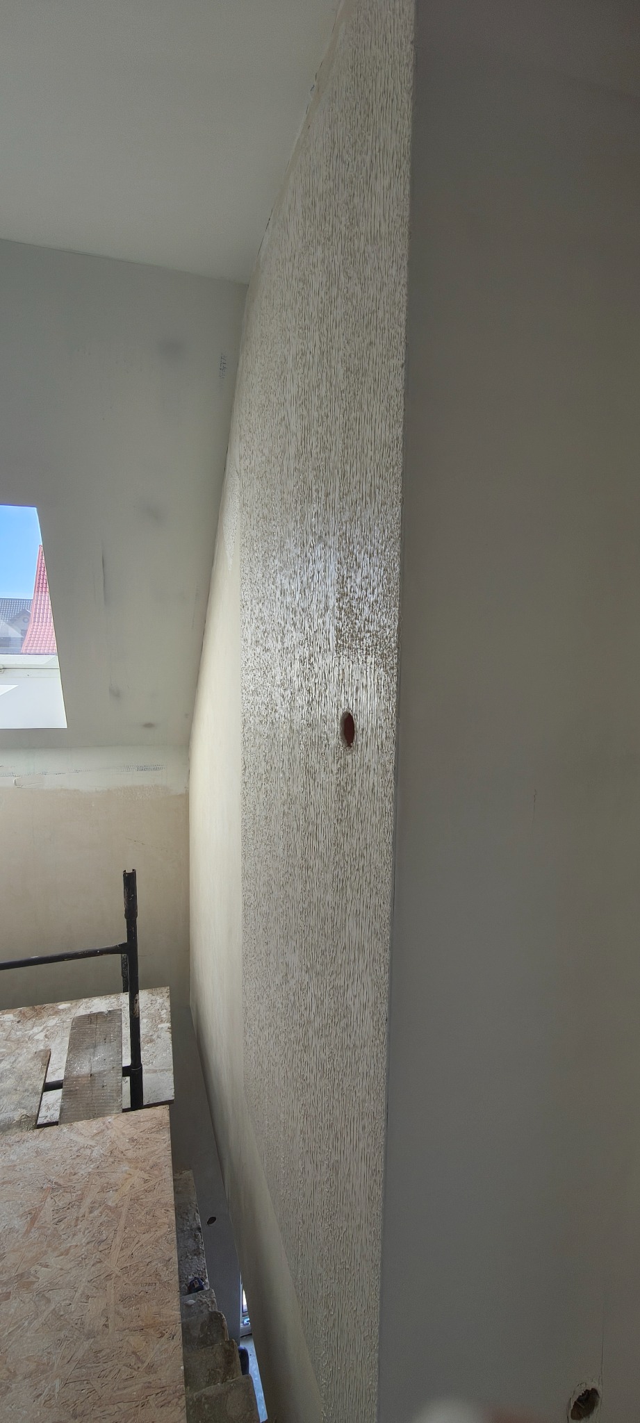Ściana po nałożeniu dekoracyjnej struktury, widoczny fragment rusztowania i okno dachowe w skosie. Teksturowana powierzchnia ściany w jasnym odcieniu beżu.
