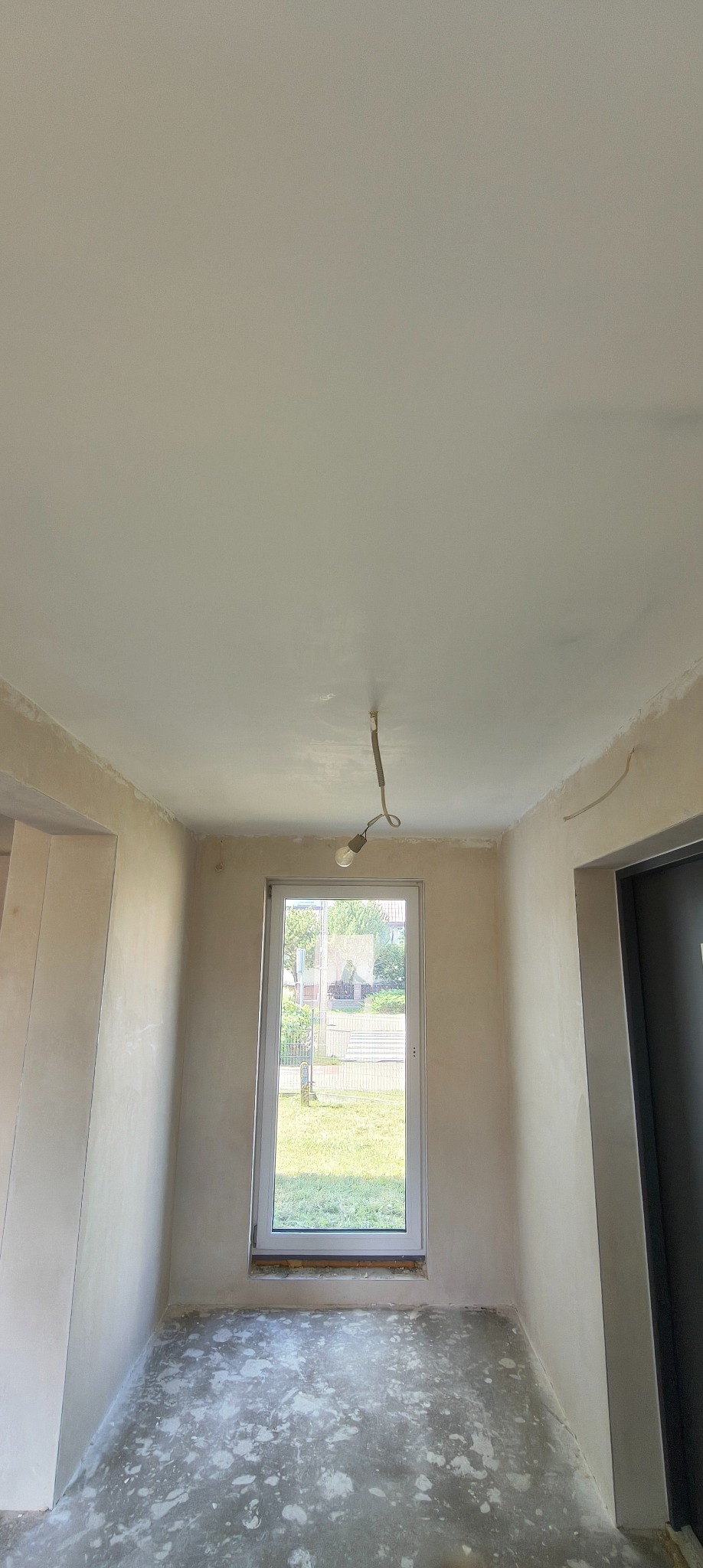 Pokój po szpachlowaniu ścian i sufitu, przygotowany do dalszych prac wykończeniowych. Widoczne okno z widokiem na zieleń oraz wisząca żarówka.