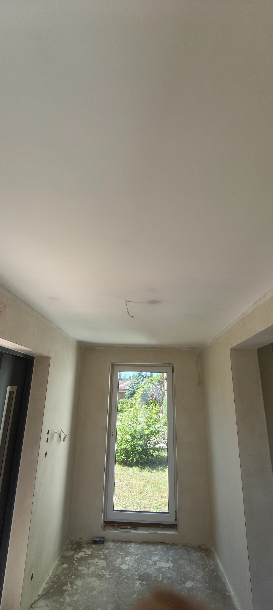 Wnętrze pokoju po szpachlowaniu ścian i sufitu, widoczne wystające kable, okno z widokiem na zieleń. Przygotowanie do dalszych prac wykończeniowych.