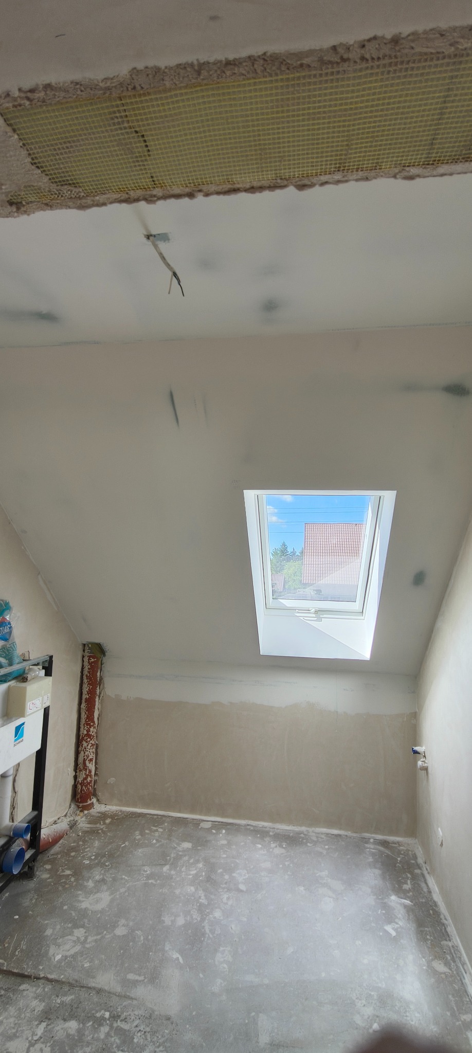 Poddasze w trakcie prac wykończeniowych: widok na fragment sufitu z siatką, ścianę połatana gipsem, okno dachowe wpuszczające światło dzienne.