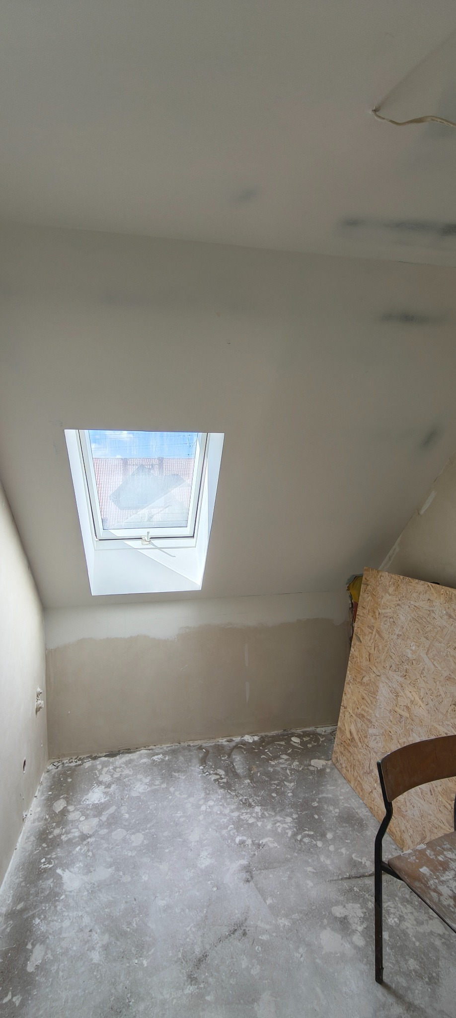 Pokój na poddaszu w trakcie prac wykończeniowych: ściany częściowo wyszpachlowane, widoczne okno dachowe i płyta OSB oparta o ścianę. Podłoga zakurzona.